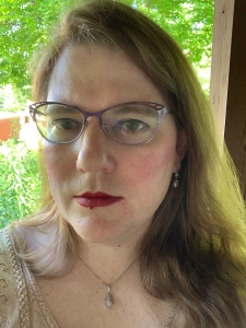 Auburn haired woman in dark red lipstick wearing two-toned purple eyeglasses, indistinct dangling earrings, and a necklace with a teardrop shaped pendant. Background is assorted indeterminate greenery.